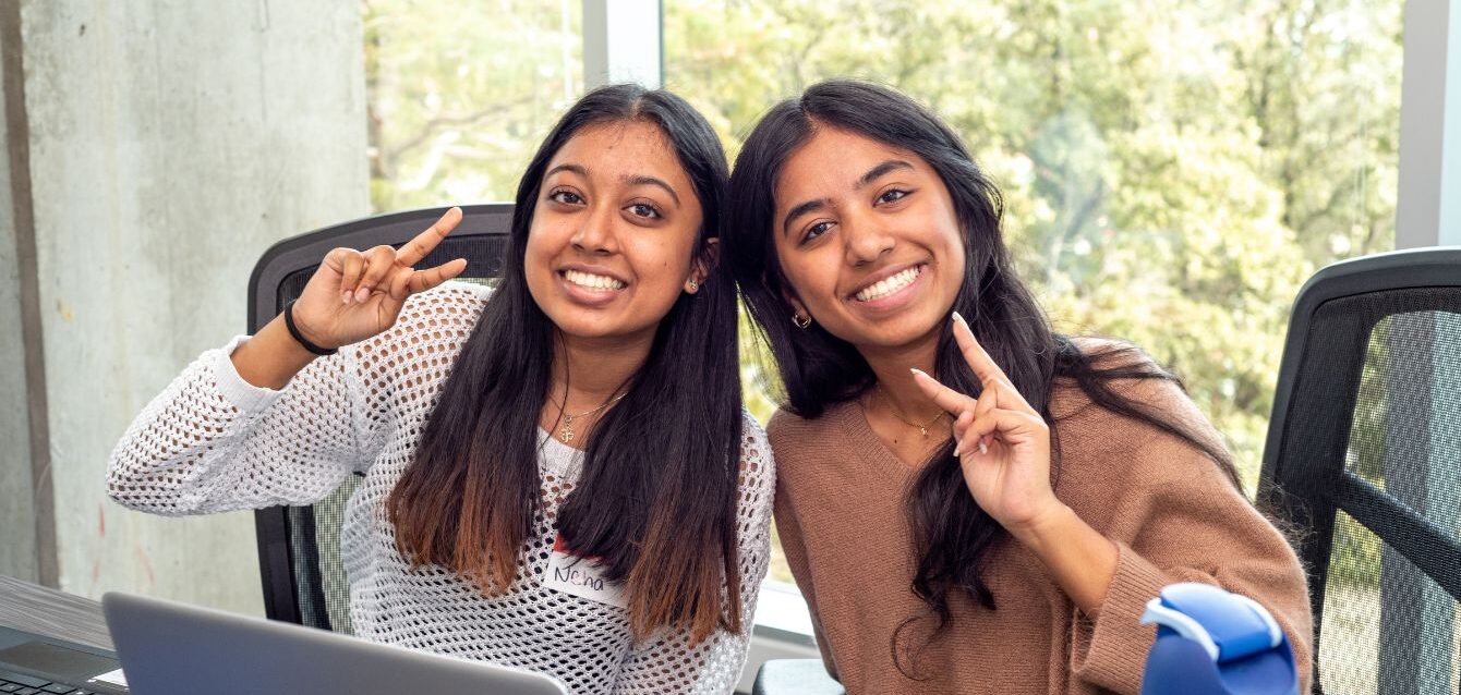 Two bootcamp students sitting side by side at a table indoors, smiling warmly at the camera and holding up peace signs with their hands