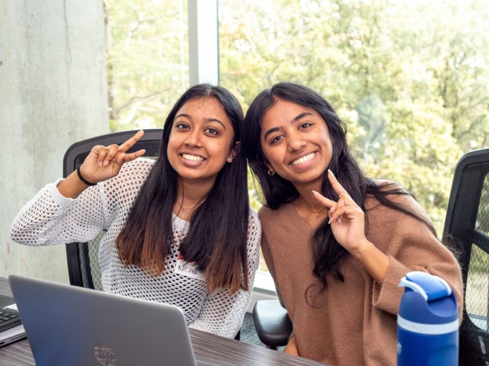 Two bootcamp students sitting side by side at a table indoors, smiling warmly at the camera and holding up peace signs with their hands